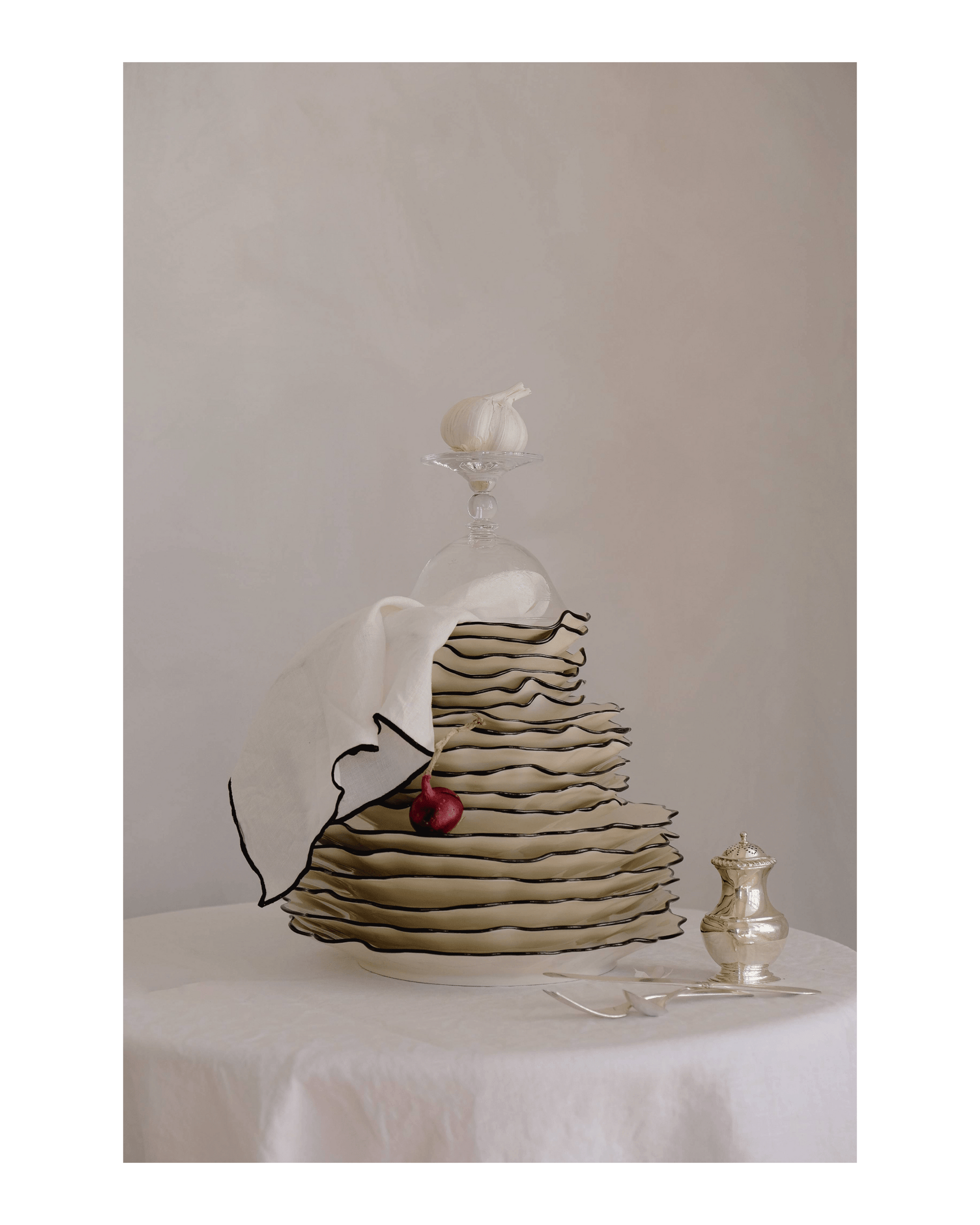 Cream coloured plates with a black edge are piled high, a red shallot and a bulb of garlic are placed on the stack. A white napkin is placed under an upside coupe glass at the top.