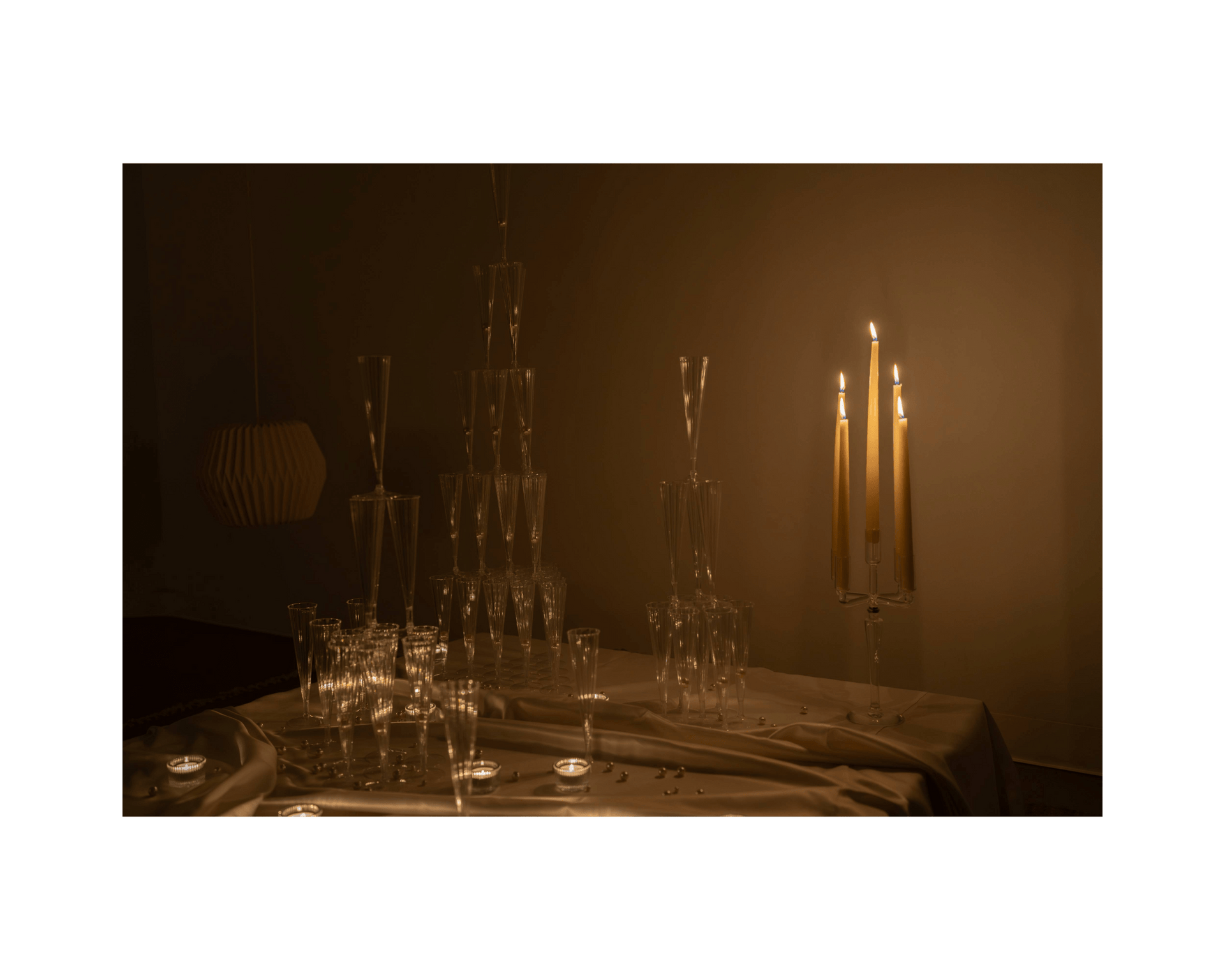 Towers of champagne glasses are stacked on a table with a satin tablecloth. A lit candelabra sits on the right side of the table.