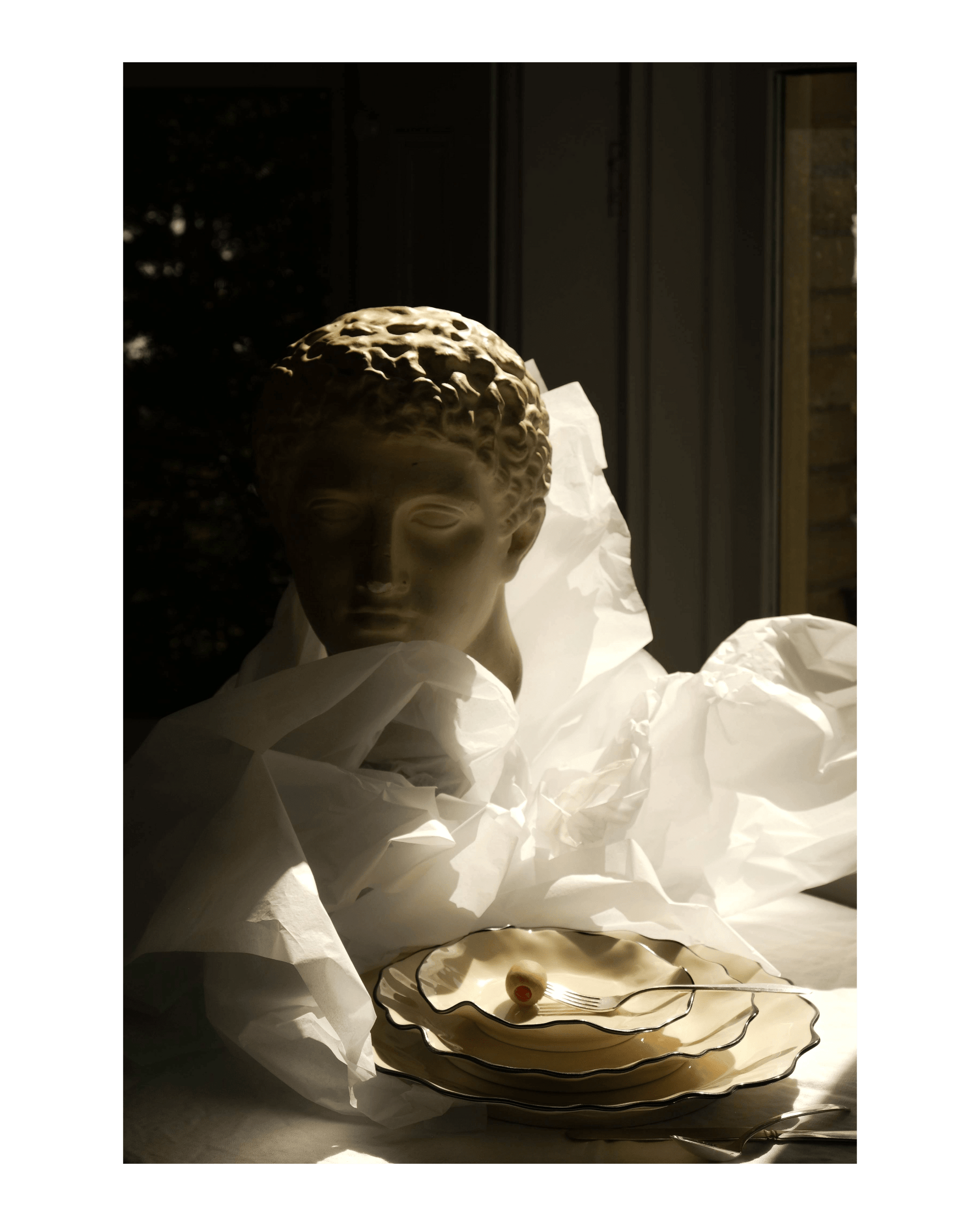A classical stone bust emerges from tissue paper behind a stack of cream plates with black detailing on the edges
