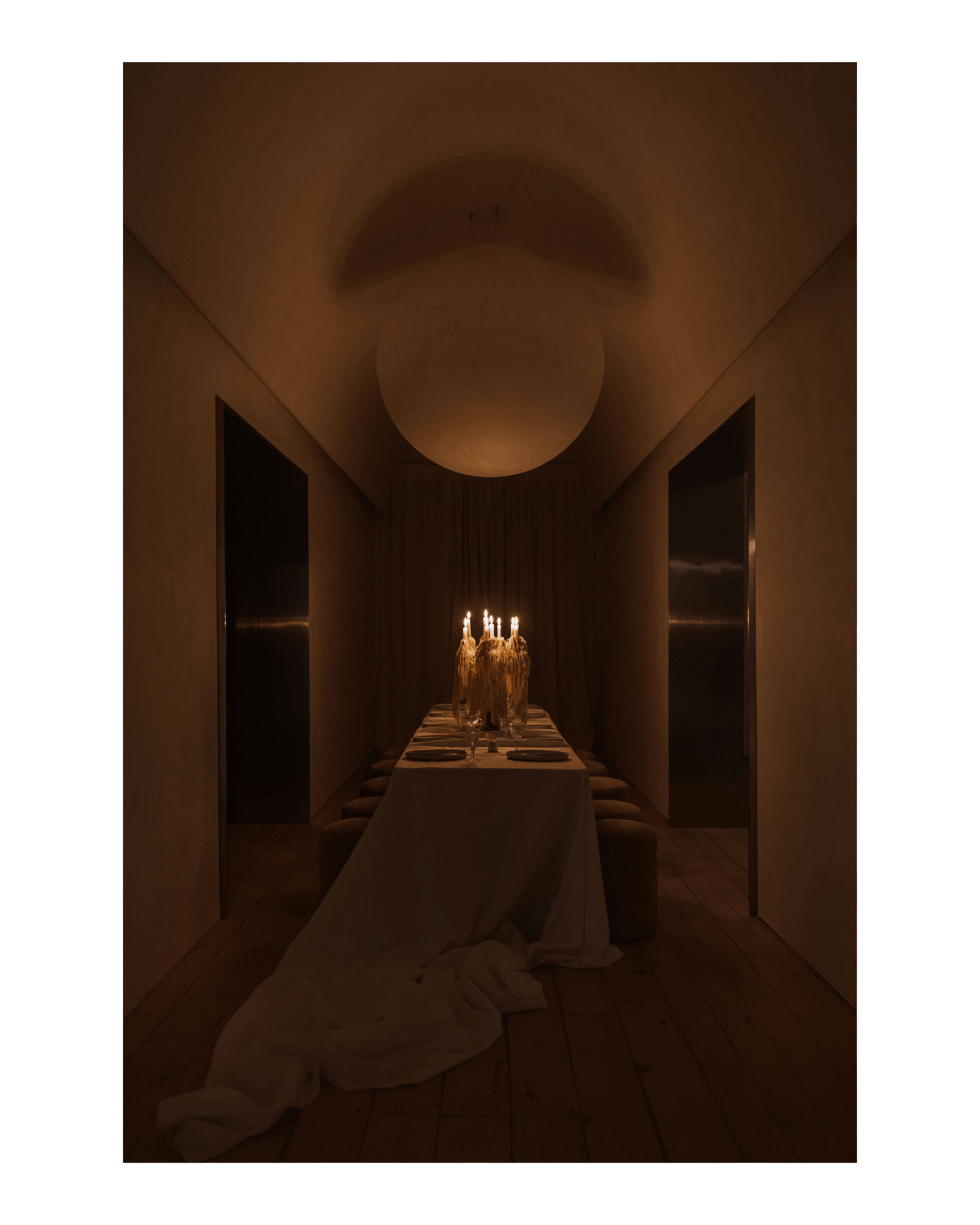 A lit candelabra sits on a dining table under a large circular pendant light. A white tablecloth spills over into a dramatic pile on the floor