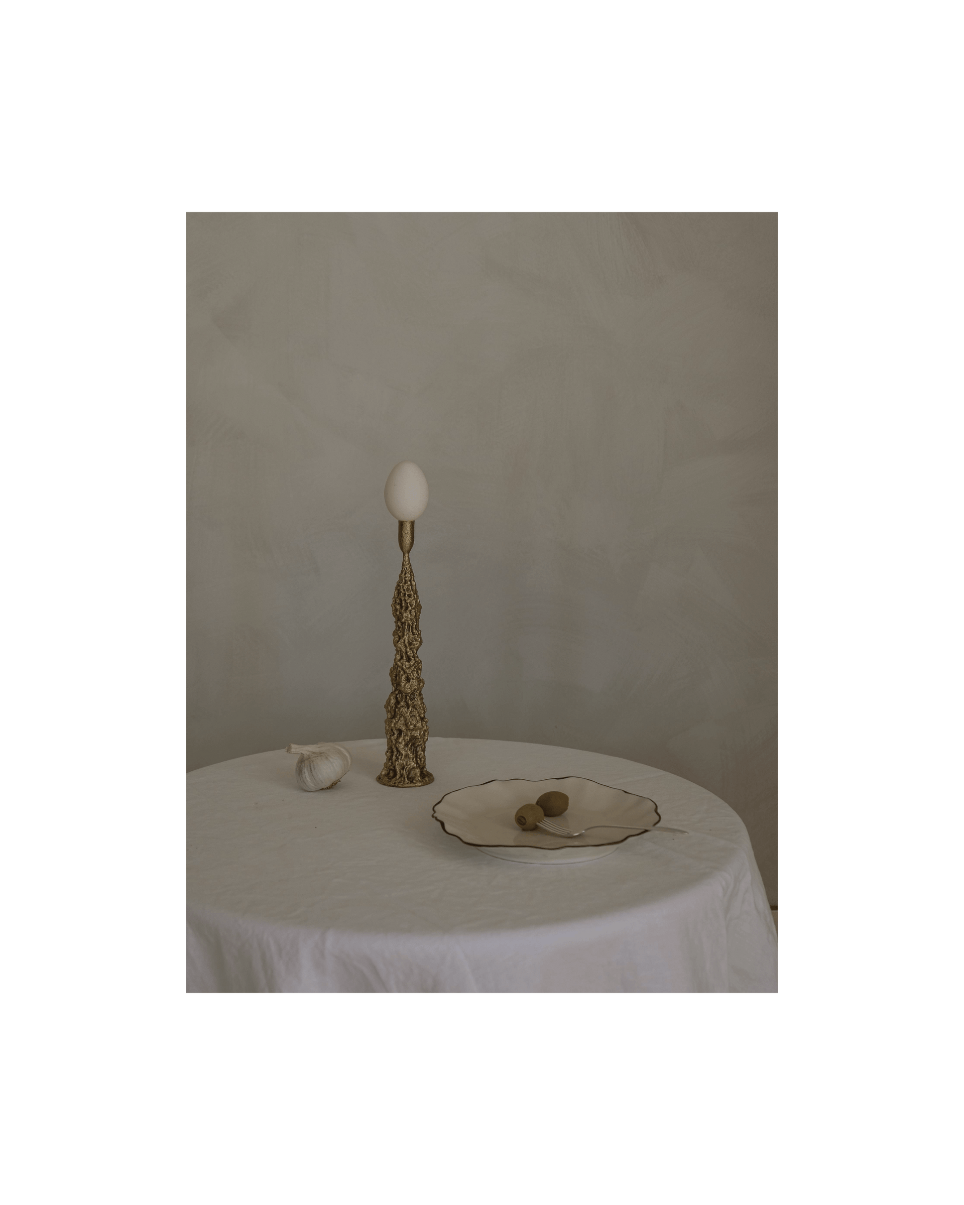 A gold decorative candlestick with an egg perched on top sits on a table. Also on the table - a bulb of garlic and a plate with two olives on it.