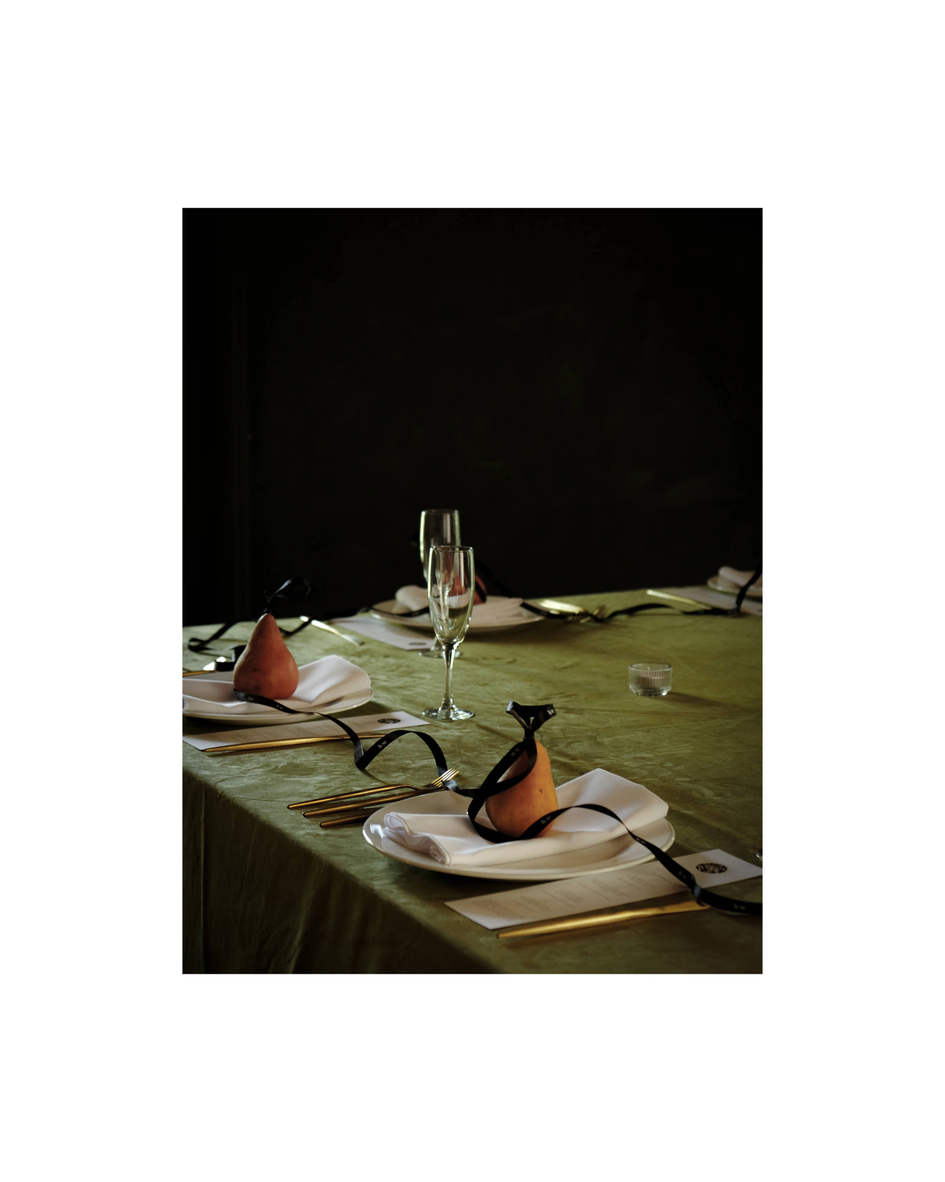 A table is set with a light green velvet tablecloth, golden brown pears sit on top of each plate. A black satin Lune branded ribbon curls across each place setting, tying the pears together
