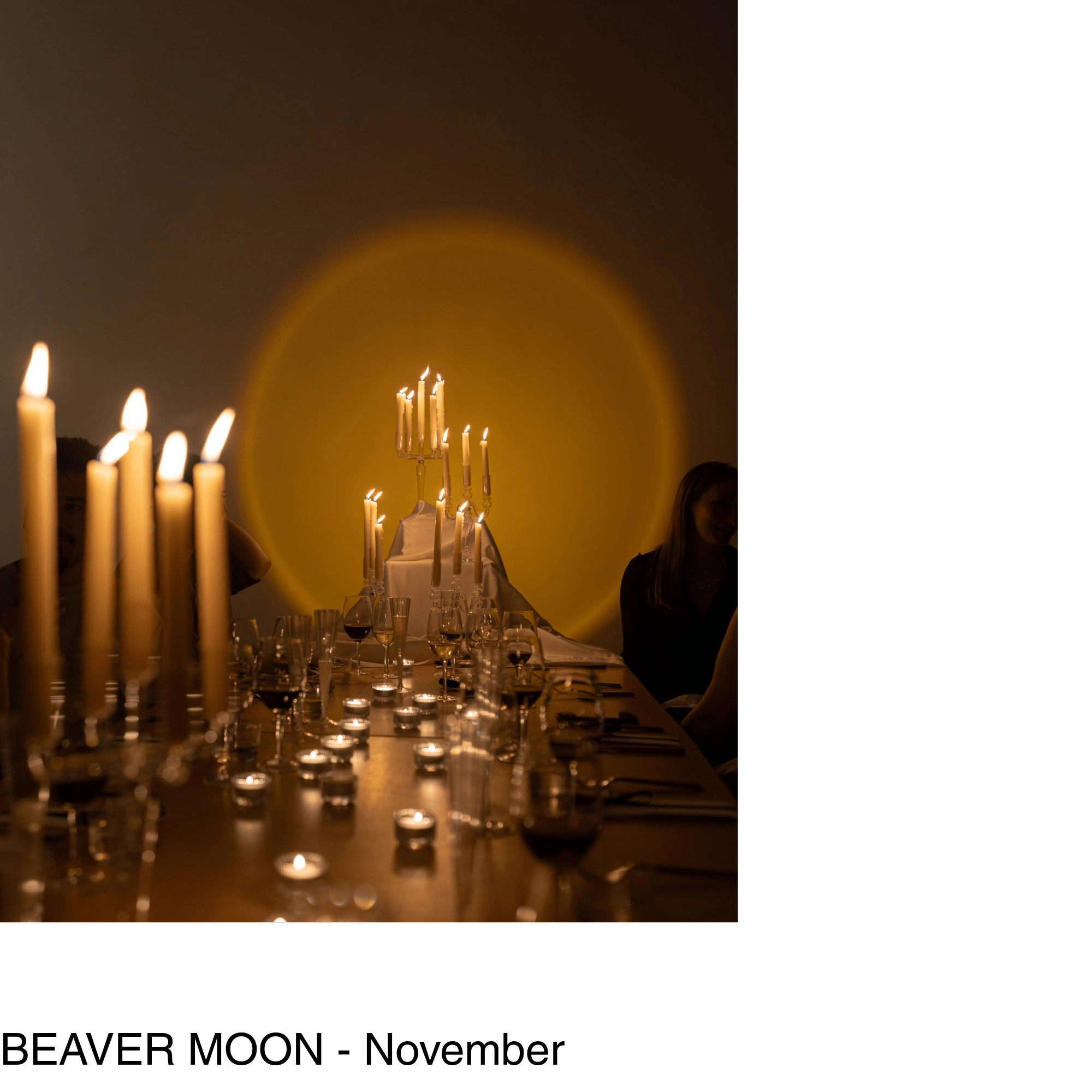 a series of lit candles, wine glasses and dishes are scattered across a long dining table - a large yellow circle of light glows on the wall behind the table.
