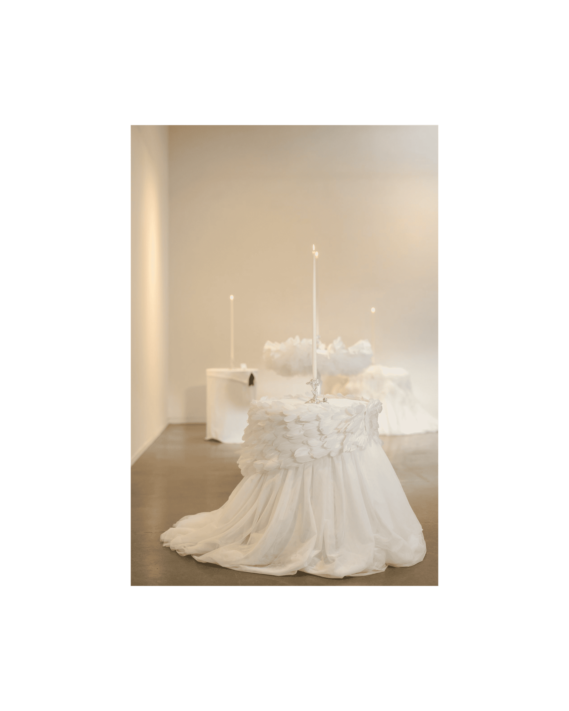 Four bespoke table cloths sit on tables in a large white gallery space, each with a tall lit candle placed in the centre of the tables. One sits in the foreground.